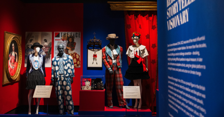 Photograph of a museum exhibition showcasing fashion designs inspired by storytelling and rebellion, featuring four mannequins dressed in bold, patterned outfits against red and blue backdrops. Display includes framed artwork, informational plaques, and a prominent blue panel with white text explaining the exhibit's theme and historical context.